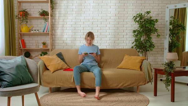 Молодая женщина проводит время дома. Молодая женщина тысячелетия сидит на диване в гостиной, печатает номер кредитной карты в приложении для смартфонов, делает покупки онлайн. Концепция повседневной жизни.