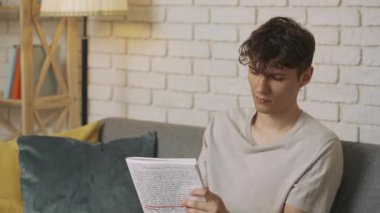 Evde okuyan genç bir öğrenci. Odaklanmış yetişkin üniversite öğrencisi gündüz vakti oturma odasında kanepede oturuyor ve ödev yapıyor, elinde defter okuma denemesi tutuyor. Eğitim kavramı.