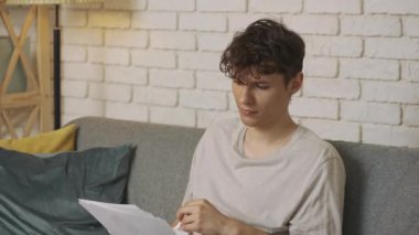 Evde okuyan genç bir öğrenci. Odaklanmış yetişkin bir üniversite öğrencisi gündüz vakti oturma odasında oturmuş ödev yapıyor, kompozisyon okuyor ve havaya kağıt çarşaf fırlatıyor. Eğitim kavramı.