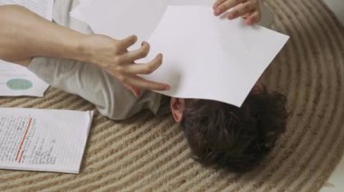 Evde okuyan genç bir öğrenci. Odaklanmış yetişkin bir üniversite öğrencisi gündüz vakti apartman dairesinde yerde yatıyor ve ödevlerini yapıyor stresten havaya kağıt çarşaf fırlatıyor. Eğitim kavramı.