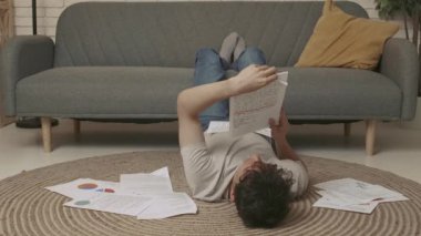 Evde okuyan genç bir öğrenci. Odaklanmış yetişkin üniversite öğrencisi gündüz vakti oturma odasında yerde uzanıyor ve ödev yapıyor. Kağıt çarşaflarla kaplı defterleri okuyor. Eğitim kavramı.
