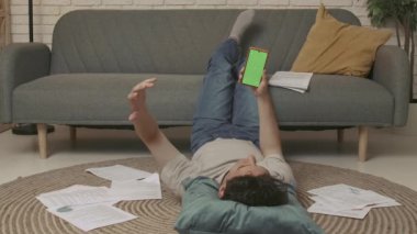 Evde okuyan genç bir öğrenci. Genç bir üniversite öğrencisi odada yerde yatıyor ve duygusal olarak yeşil ekranlı akıllı bir telefonla konuşuyor. Eğitim kavramı.