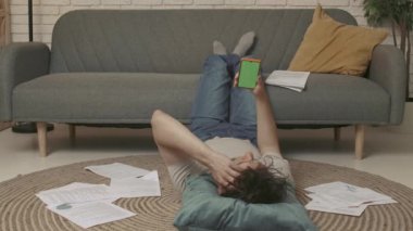 Evde okuyan genç bir öğrenci. Genç bir üniversite öğrencisi odada yerde yatıyor ve duygusal olarak yeşil ekranlı akıllı bir telefonla konuşuyor. Eğitim kavramı.