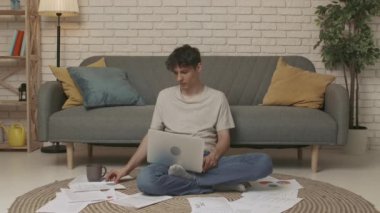 Evde okuyan genç bir öğrenci. Odaklanmış yetişkin üniversite öğrencisi gündüz vakti apartman dairesinde yerde oturuyor ve dizüstü bilgisayarda ödevlerini yapıyor. Eğitim kavramı.