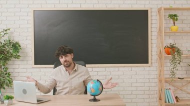 Erkek bir öğretmen, arka planda tahtası olan bir sınıfta coğrafya dersi veriyor. Adam öğrencilere bilgi veriyor ve bir küreyi işaret ediyor. Eğitim kavramı.