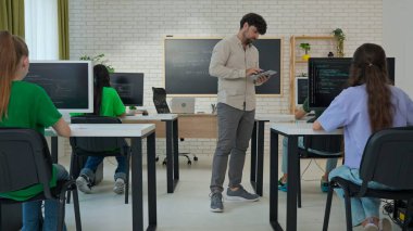 Eğitmen, modern ve iyi donanımlı bir sınıfta öğrencilerin bilgisayarlarındaki kodlama projelerini aktif olarak izlemek ve onlara yardımcı olmak için bir tablet kullanır..