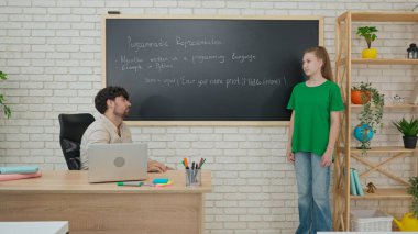 Modern bir sınıfta bir öğrenci öğretmeninin yanında durur ve tahtaya yazılmış bir Python programlama örneğini tartışır..