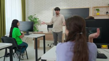 Bir bilgisayar bilimleri ve programlama eğitmeni bir soru sorar ve öğrenciler cevap vermek için ellerini kaldırır. Modern sınıf bir karatahta ve bilgisayarlarla donatılmıştır..