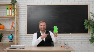 Okul öğretmeni sınıfta gençlerle konuşuyor. Sınıftaki masada oturan erkek üniversite öğretmeni elinde krom renkli yeşil ekran modeli, işaret parmağı olan akıllı telefon tutuyor. Eğitim kavramı.