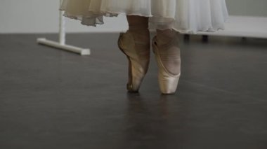 Beyaz elbiseli çıplak bir balerin, beyaz güneşli bir odada dans ediyor. Siyah bir zemin de bir bale barının yanında.