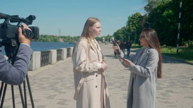 Bir TV muhabiri şehir manzaralı bir rıhtımda genç bir kadınla röportaj yapıyor. Kamera operatörü etkileşimi yakalar ve haber bölümüne dinamik bir katman ekler..
