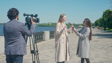 Bir TV muhabiri şehir manzaralı bir rıhtımda genç bir kadınla röportaj yapıyor. Kamera operatörü etkileşimi yakalar ve haber bölümüne dinamik bir katman ekler..