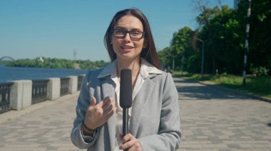 Nehir kenarında canlı bir günde yakalanan erkek muhabir, elinde mikrofonuyla canlı bir haber bölümü sunuyor. İş elbiseli bir kadın doğrudan kameraya bakarken haberleri sunuyor..