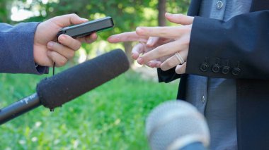 Interview outside in a park. A man answers questions from reporters. Hands with a microphone, a smartphone and a voice recorder are visible in front of him recording the interview. clipart