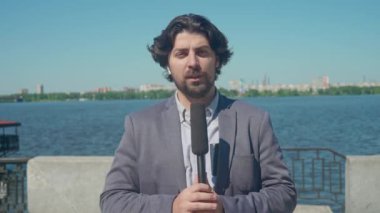 Nehir kenarında canlı bir günde yakalanan erkek muhabir, elinde mikrofonuyla canlı bir haber bölümü sunuyor. İş elbisesi giymiş orta yaşlı bir adam doğrudan kameraya bakarken haberleri iletir..