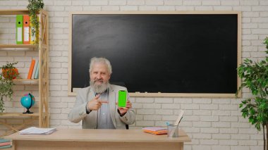 Ders sırasında oditoryumda öğretmen. Yaşı ilerlemiş üniversite profesörü sınıfın masasına oturmuş elinde akıllı telefon kroma anahtar yeşil ekran ile onu işaret ediyor. Eğitim kavramı.
