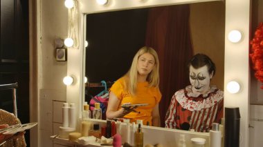 Makyöz soyunma odasında Cadılar Bayramı 'na hazırlanan bir adama palyaço makyajı yapıyor. Sanatçı yüzü detaylandırmaya odaklanırken, çeşitli makyaj araçları ve ürünleri masanın etrafına saçıldı