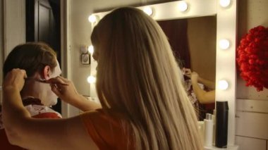 Makyöz soyunma odasında Cadılar Bayramı 'na hazırlanan bir adama palyaço makyajı yapıyor. Sanatçı yüzü detaylandırmaya odaklanırken, çeşitli makyaj araçları ve ürünleri masanın etrafına saçıldı