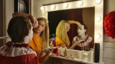 Makyöz soyunma odasında Cadılar Bayramı 'na hazırlanan bir adama palyaço makyajı yapıyor. Sanatçı yüzü detaylandırmaya odaklanırken, çeşitli makyaj araçları ve ürünleri masanın etrafına saçıldı