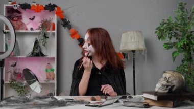 Kadın nüfuzlu kişi canlı video makyaj dersi veriyor. Evde pelerinli genç bir kadın güzellik blogcusu Cadılar Bayramı partisi makyajı için video dersleri veriyor, kamerayla konuşuyor, cadı makyajını fırçayla onarıyor..