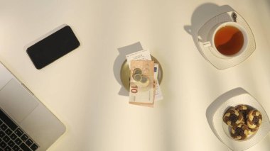 Bir kafede ödenmiş bir faturanın üst görüntüsü. Tabağın üzerinde çek, bozuk para ve avro banknotları var. Masanın üzerinde de yarı sarhoş bir fincan çay ve bir dizüstü bilgisayar var. Para ve ödeme kavramı