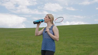 Genç aktif bir kadın açık hava badminton oyunu sırasında su içmek için mola verir. Elinde bir su şişesi ve raket parçalı bulutlu bir gökyüzünün altında geniş yeşil bir alanda..