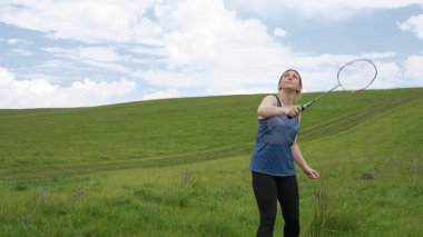 Genç bir kadın mavi gökyüzünün altındaki geniş bir çimenlikte tek başına badminton oynuyor. Spor, sağlık ve doğanın güzelliği için mükemmel bir tasvir..