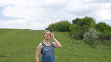 Düşünceli genç bir kadın, etek giymiş, yemyeşil bir alanda duruyor, uçsuz bucaksız gökyüzünün altındaki mesafeye bakıyor..