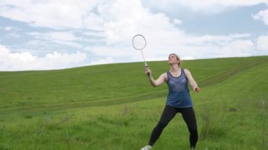 Genç bir kadın mavi gökyüzünün altındaki geniş bir çimenlikte tek başına badminton oynuyor. Spor, sağlık ve doğanın güzelliğini tasvir etmek için mükemmel. Yavaş çekim.