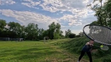 İki arkadaş, dinamik bir gökyüzünün altındaki geniş yeşil bir alanda gündelik badminton oynamaktan zevk alıyor. Bir kadın ve bir erkek aktif olarak badminton oynuyor ve raketlerle bir mekik horozuna vuruyorlar. Yavaş çekim.