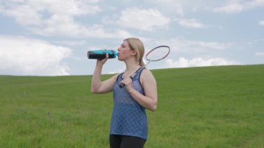 Genç aktif bir kadın, açık hava badminton oyunu sırasında su içmek için ara verir. Parçalı bulutlu bir gökyüzünün altında, geniş yeşil bir alanda su şişesi ve raket tutar. Yavaş çekim.