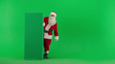 Noel ve Yeni Yıl yaratıcı konsepti. Krom anahtar yeşil ekranda Noel Baba kılığında bir adam portresi. Kırmızı kıyafetli Noel Baba boş tahtanın arkasında duruyor ve onu işaret ediyor..