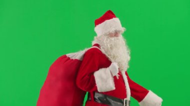 Noel ve Yeni Yıl yaratıcı konsepti. Krom anahtar yeşil ekranda Noel Baba kılığında bir adam portresi. Kırmızı kıyafetli Noel Baba hediye çantası taşıyor ve yürüyor. Şut. Yan görünüm