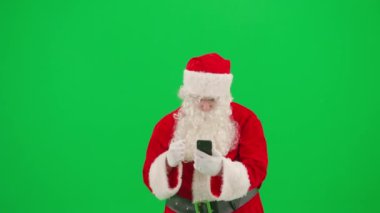 Noel ve Yeni Yıl yaratıcı konsepti. Krom anahtar yeşil ekranda Noel Baba kılığında bir adam portresi. Noel Baba akıllı telefondan spor maçı izliyor, mutlu kazanma ifadesi.
