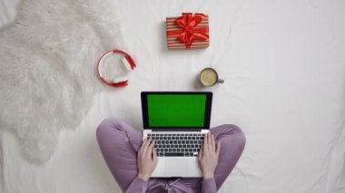 Tatil, tatil ve online reklam alışverişi. Yatakta hediye kutusu ve kahve kupası olan bir kızın üst görüntüsü alet krom anahtar yeşil ekran kullanıyor. Yatakta pijamalı bir kadın dizüstü bilgisayar tutuyor..