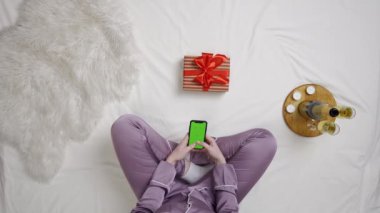 Tatil, tatil ve online reklam alışverişi. Yatakta hediye kutulu ve şampanya kadehli bir kızın üst görüntüsü krom anahtar yeşil ekran kullanıyor. Yatakta akıllı telefondan mesaj atan bir kadın..