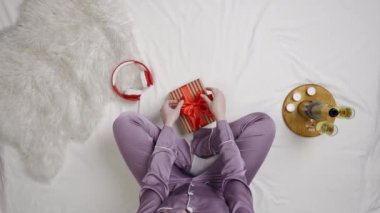 Tatil, tatil ve online reklam alışverişi. Yatakta hediye kutusu, kulaklığı ve bir şişe şampanyası olan kız manzarası. Pijamalı kadın yatağa oturur ve hediye kutusunu kırmızı kurdeleyle sarar..
