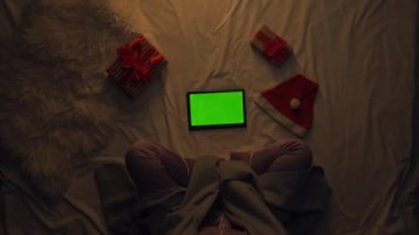 Tatil boş zaman konsepti. Battaniyeyle kaplı yatağın üzerindeki kızın üst görüntüsü. Geceleri pijamalı bir kadın yatakta oturmuş televizyon izliyor ve krom renkli yeşil ekran arka planı olan tablet kullanıyor..