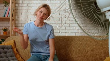 Genç bir bayan evde vakit geçiriyor. Genç milenyum kadını oturma odasındaki koltukta oturmuş, havalandırma vantilatörünün altında dinleniyor, temiz hava üflüyor, yakın çekim. Sıkıcılık günlük yaşam konsepti.
