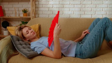 Young female spending time at home. Young millennial woman laying on the sofa in the living room waving fan, coping with summer heat, close up shot. Stuffiness everyday life concept. clipart