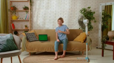 Young female spending time at home. Young millennial woman sitting on the sofa in the living room under electric fan, laptop with chroma key green screen on the couch. Stuffiness everyday life concept clipart