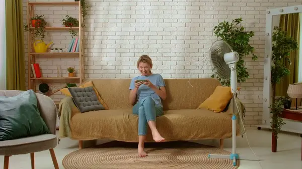 stock image Young female spending time at home. Young millennial woman sitting on the sofa in the living room resting under electric fan, playing video game on smartphone. Stuffiness everyday life concept.