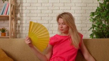 Genç bir bayan evde vakit geçiriyor. Genç milenyum kadını oturma odasındaki kanepede oturmuş, eğlenceli bir şekilde yelpaze sallıyor, yaz sıcağıyla başa çıkıyor, yakın çekim yapıyor. Sıkıcılık günlük yaşam konsepti.