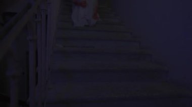 Genç bir kadın karanlıkta merdivenlerde. Beyaz elbiseli ürkütücü hayalet kadın karanlık perili evde merdivenlerde oturuyor, korkunç maskeli kırmızı balonla saklanıyor. Cadılar Bayramı korku reklam konsepti.