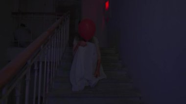 Genç bir kadın karanlıkta merdivenlerde. Beyaz elbiseli ürkütücü hayalet kadın karanlık perili evde merdivenlerde oturuyor, korkunç maskeli kırmızı balonla saklanıyor. Cadılar Bayramı korku reklam konsepti.