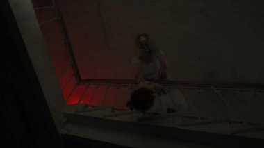 Genç bir kadın karanlıkta merdivenlerde. Beyaz elbiseli ve korkunç maskeli ürkütücü hayalet kadın karanlık perili evde merdivenlerde oyuncak tutarken tırabzanların üzerinden eğiliyor. Cadılar Bayramı korku reklam konsepti.