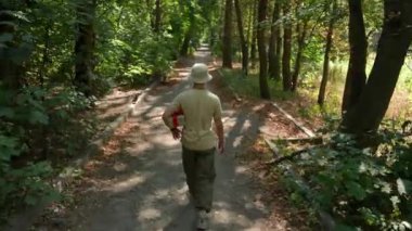 Genç bir adam elinde bir hediyeyle parkta yürüyor. Güneşli bir yaz gününde, günlük giysiler içinde park yolunda yürüyen bir adamın arka görüntüsü. Tatil ve dinlenme konsepti.