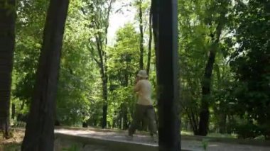 Genç bir adam telefonda konuşurken parkta yürür vaziyette. Güneşli bir yaz gününde, günlük giysiler içinde park yolunda yürüyen bir adamın arka görüntüsü. Eğlence ve aktif yaşam tarzı kavramı.