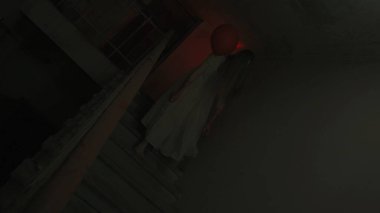 Genç bir kadın karanlıkta merdivenlerde. Beyaz elbiseli ve korkunç makyajlı ürkütücü hayalet kadın elinde kırmızı balonla karanlık perili bir evde tek başına merdivenlerde yürüyor. Cadılar Bayramı korku reklam konsepti.