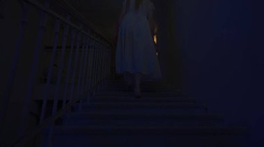 Genç bir kadın karanlıkta merdivenlerde. Beyaz elbiseli ve korkunç makyajlı ürkütücü hayalet kadın karanlık perili evde merdivenlerde tek başına yürüyor. Cadılar Bayramı korku reklam konsepti.